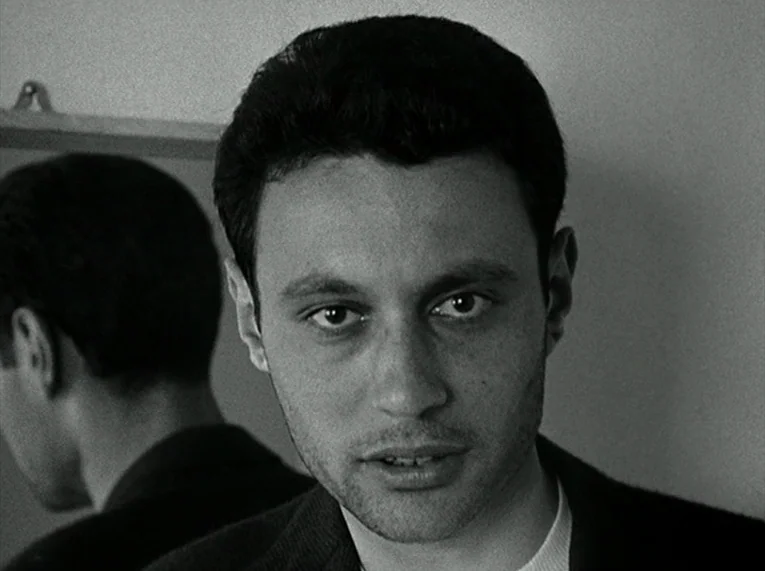 Le Petit Soldat (1963) movie still: closeup — A man stands in front of a mirror inside a spare interior room. The reflection of his head and…