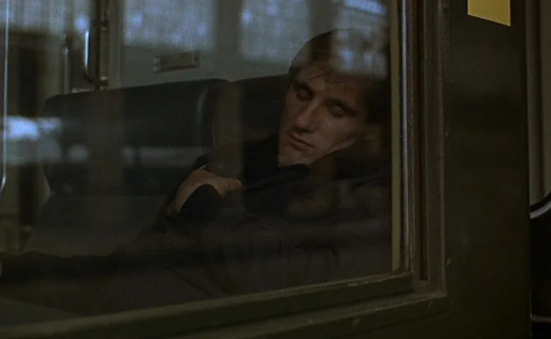 Maîtresse (1975) movie still: medium — A dark train cabin features leather seating and reflections on the window glass. A young man with dark hair…