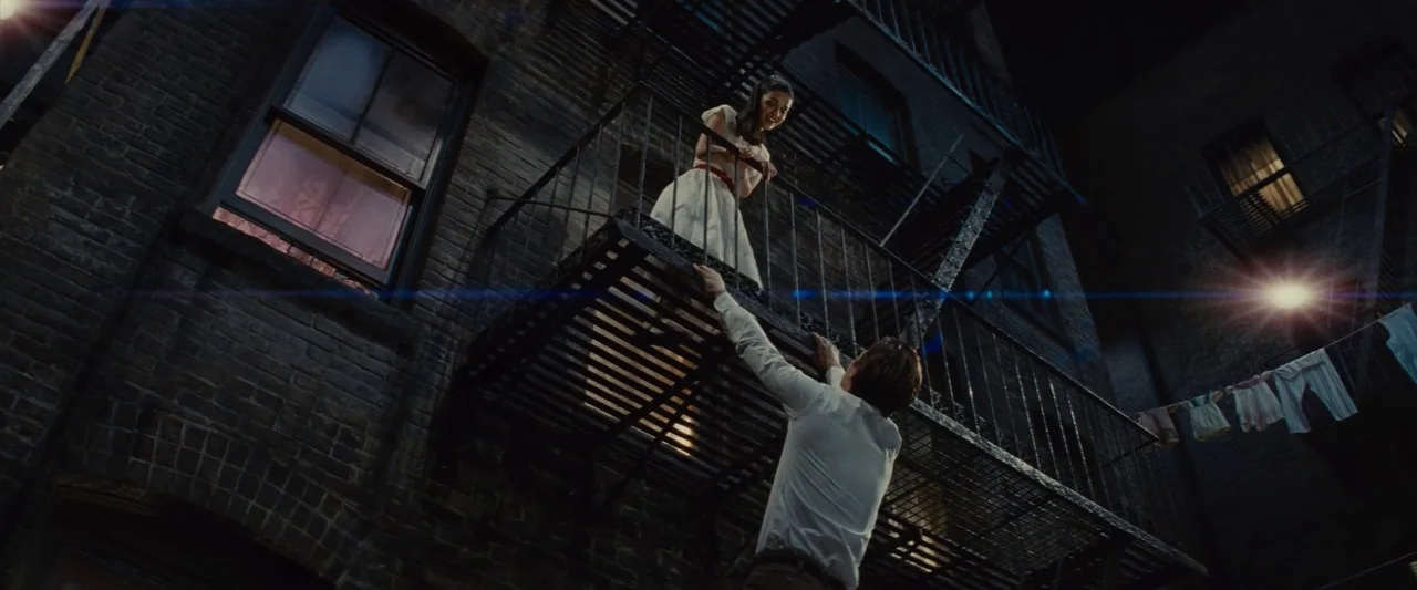 Making of: West Side Story (2021) movie still: wide — An exterior nighttime view shows a brick building wall with a rusted metal fire escape. A…