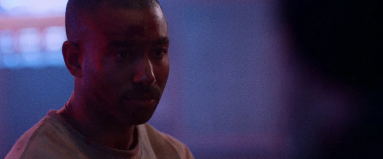 VFW movie still: closeup — A dimly lit interior space is cast in hues of blue and purple neon. A man with a buzzed haircut gazes toward the side of…