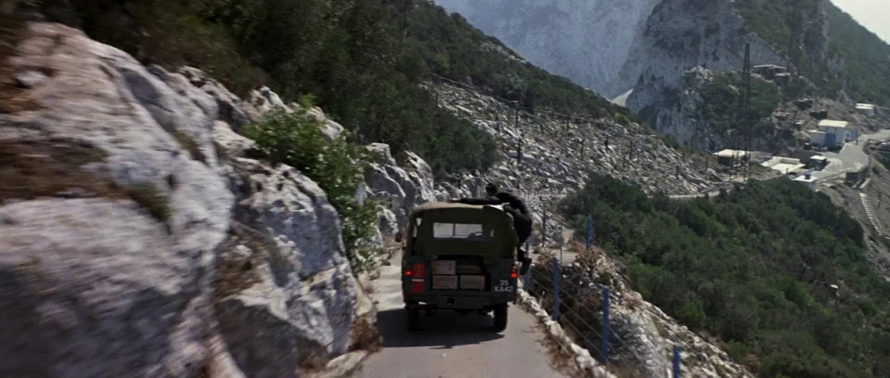 The Living Daylights (1987) movie still: wide — A narrow paved road winds along a steep white marble quarry on a mountainside. A person clings to…