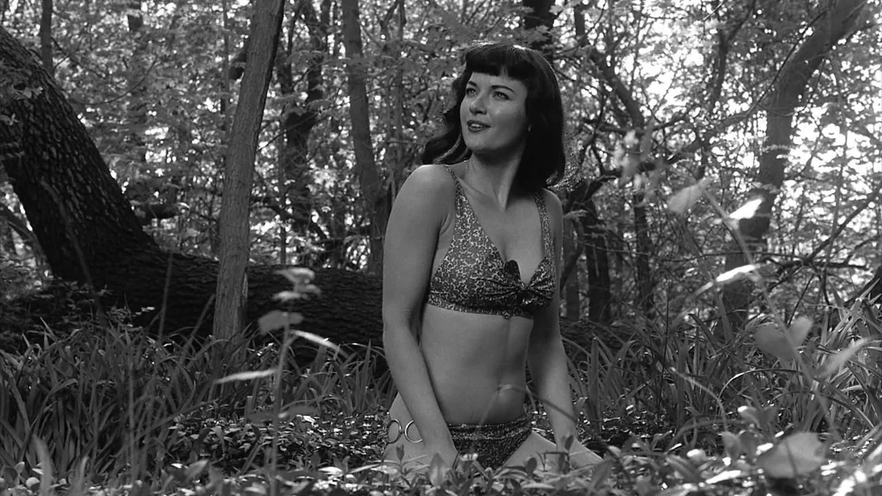 The Notorious Bettie Page (2005) movie still: medium — A dense woodland area with thick foliage and a fallen tree trunk in background. A woman in a…