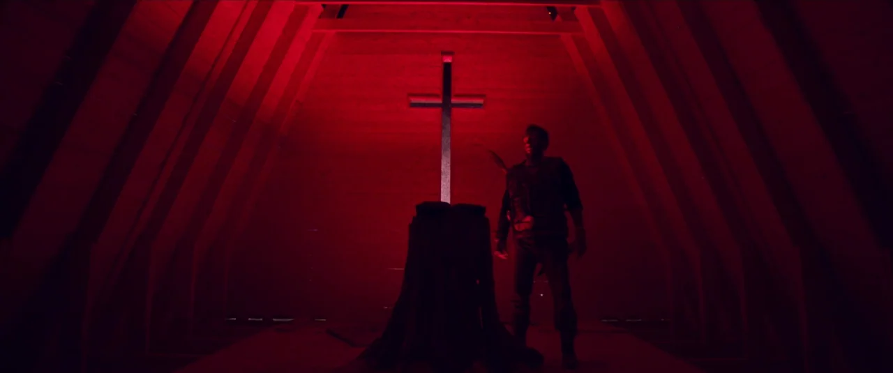 Mandy (2018) movie still: wide — An A-frame wooden structure features a large cross centered against the back wall. A man stands near a stump…