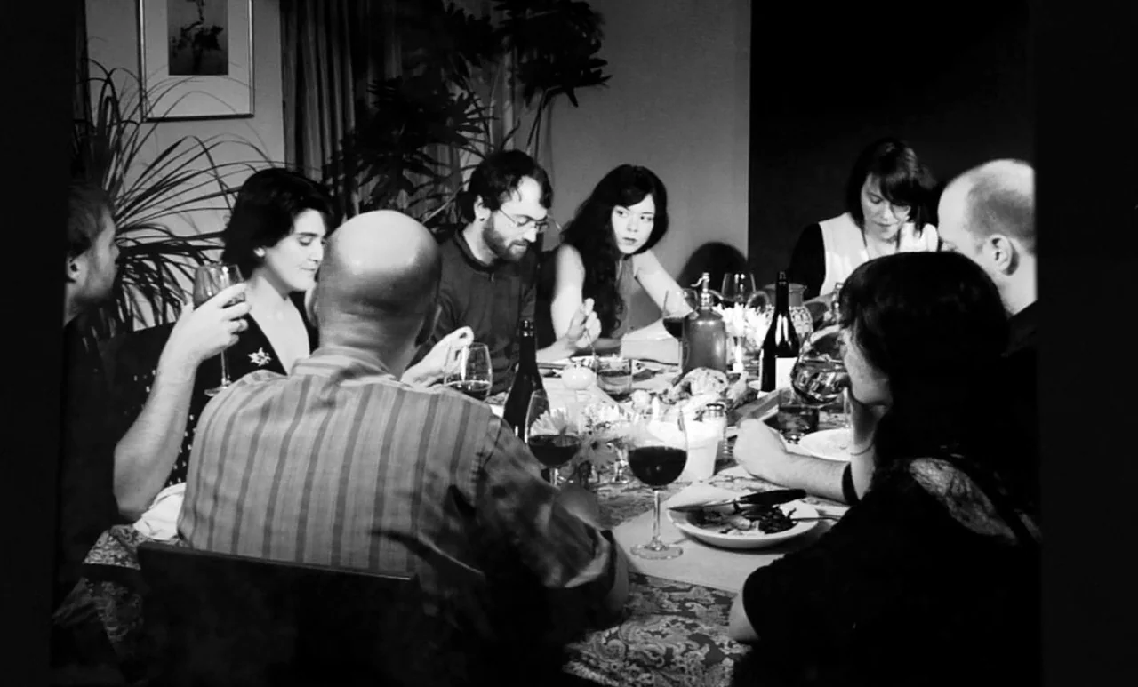 Go Down Death movie still: medium — A group of adults sits around a dining table covered with dishes and wine. They are engaged in conversation…