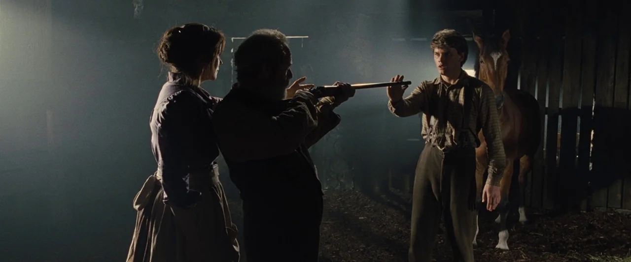 War Horse (2011) movie still: fullbody — A dark wooden barn contains three figures standing in dusty beams of light. A man points a long firearm at…