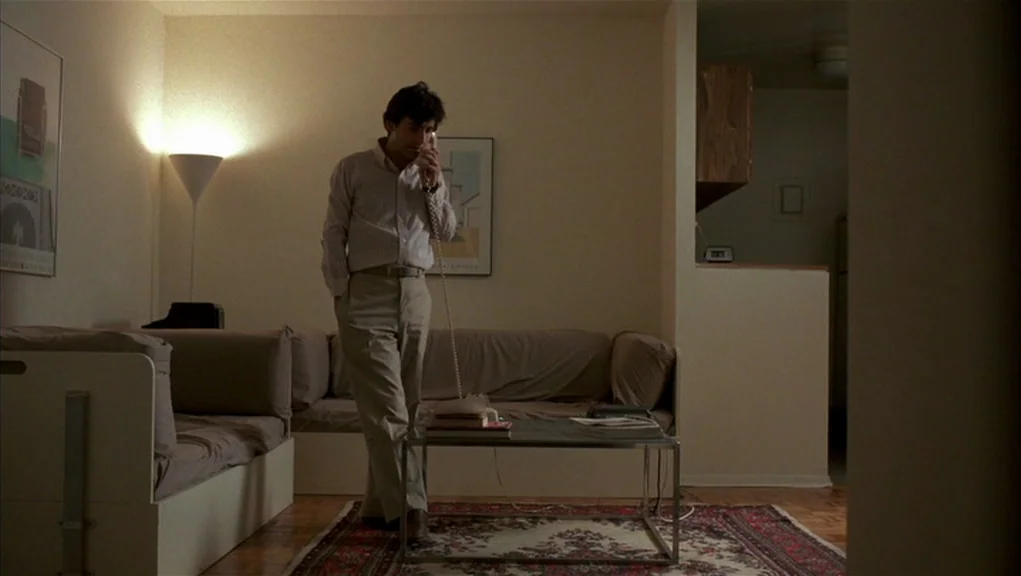 After Hours (1985) movie still: fullbody — A spartan apartment interior features a modern coffee table and a beige sofa. A man stands in the center…