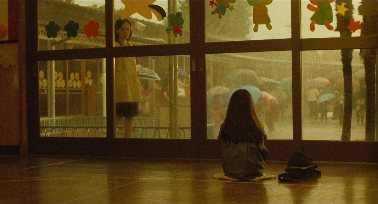 Dark Water (2002) movie still: medium — A dark indoor hallway features a large glass wall covered in paper cutouts. A child sits on the floor inside…