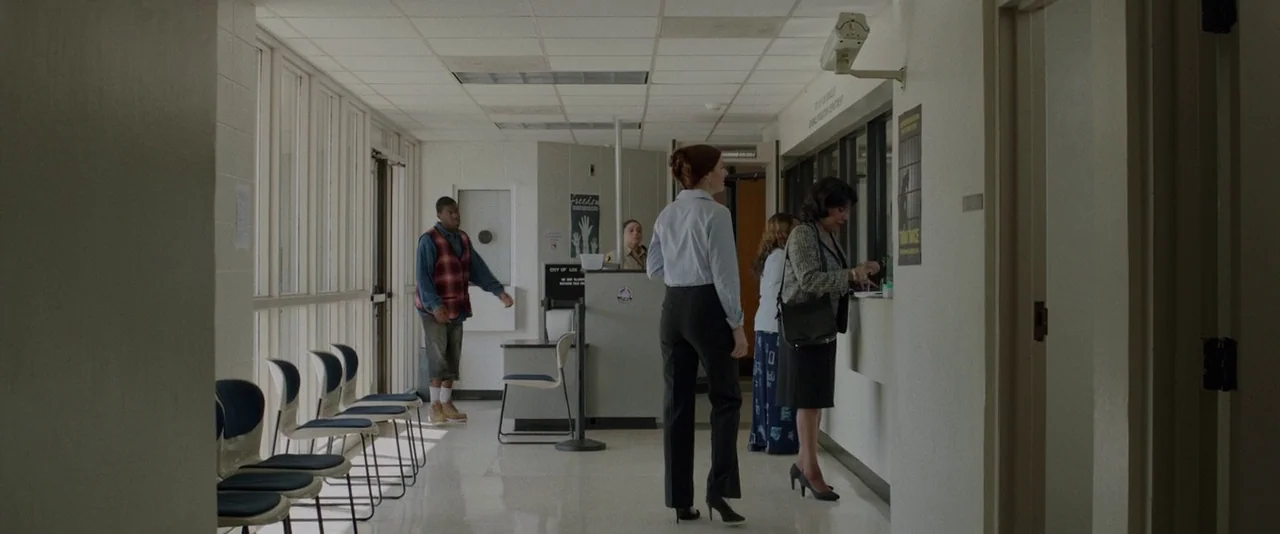 Creed (2015) movie still: wide — An institutional waiting area with beige block walls and a drop ceiling. Several people stand in a line at a…