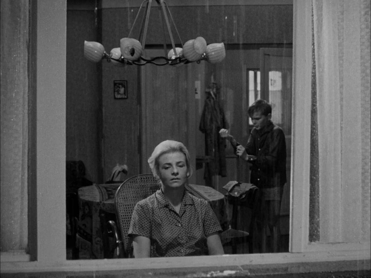 The Sun In A Net (1963) movie still: medium — A view through a window into a sparsely decorated 1960s dining space. A woman sits in the foreground…