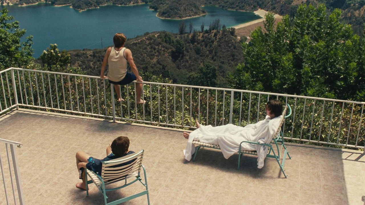 Love & Mercy (2014) movie still: wide — A spacious tiled balcony features a white metal railing overlooking a calm reservoir. Three young men lounge…