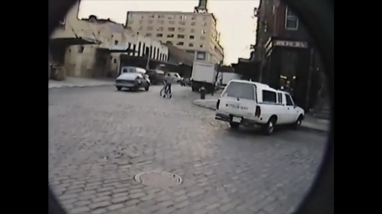 The Stroll (2023) movie still: wide — A quiet cobblestone street scene is captured through a rounded, vignetted camera lens. A white pickup truck is…