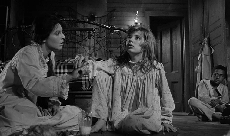 The Miracle Worker (1962) movie still: medium — A dimly lit room with wooden walls and a metal bed frame in the back. An adult woman sits with two…