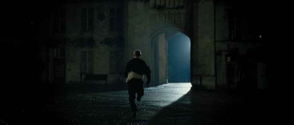 Hot Fuzz (2007) movie still: fullbody — An outdoor paved square at night framed by stone walls and a stone archway. A man in dark clothing runs…