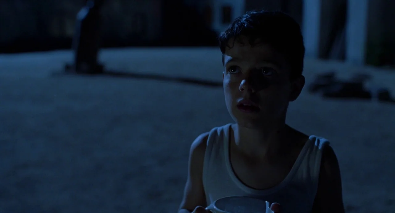 The Devils Backbone (2001) movie still: medium — A dark outdoor courtyard at night features sand on the ground and vague structures. A young boy in…