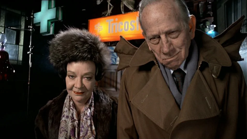 The Tenant (1976) movie still: medium — A dimly lit pharmacy interior features bright orange and green neon pharmaceutical signage background. An…