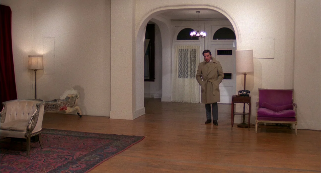 Opening Night (1977) movie still: wide — A spacious home foyer with hardwood floors features an arched doorway and sparse furniture. A man in a…