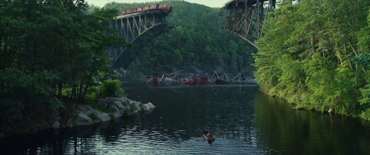 The Man Who Killed Hitler and Then the Bigfoot (2018) movie still: wide — A dark river flows through a dense forest toward a destroyed metal railway…