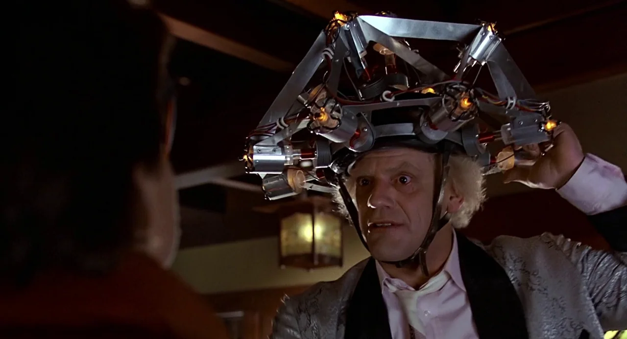 Back to the Future (1985) movie still: medium — A cluttered laboratory setting features an older man wearing a complex metal apparatus. The man…