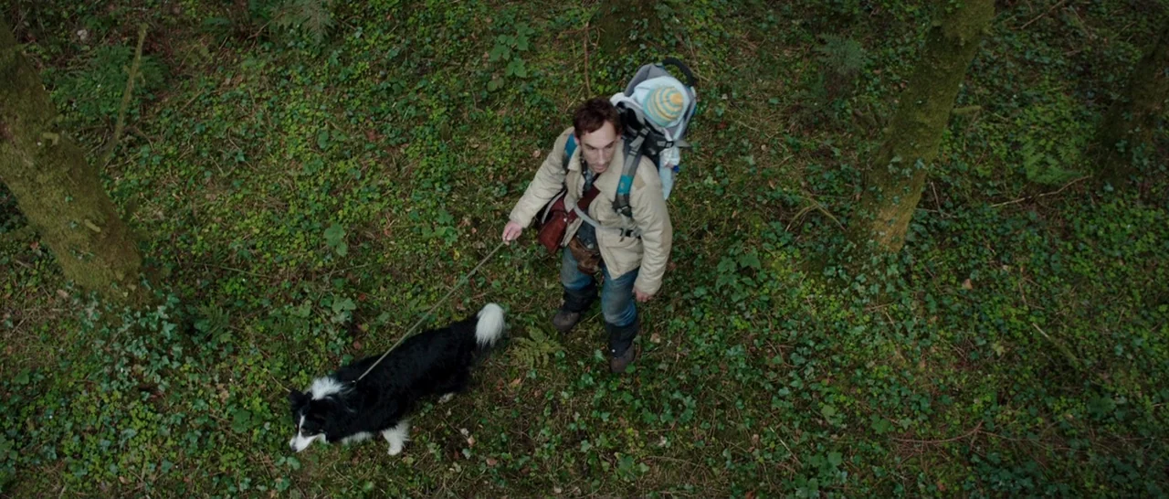The Hallow (2015) movie still: wide — A man carries an infant in a backpack while walking his dog in dense woods. The ground is carpeted with green…