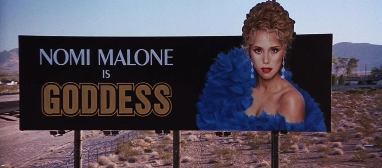 Showgirls (1995) movie still: wide — A roadside billboard features text and a portrait of a woman in the desert. The landscape is arid with sparse…