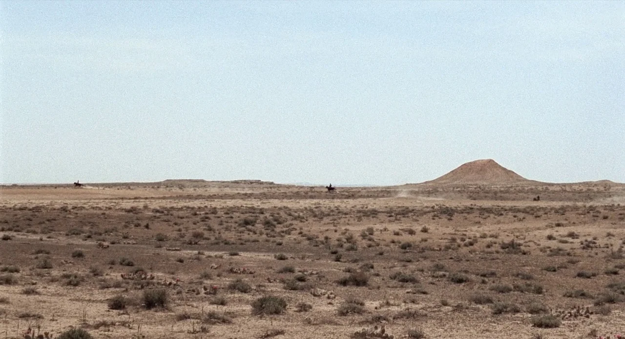 The Shooting (1966) movie still: wide — An arid, sprawling desert plain features sparse scrub brush under a clear pale sky. Three small figures on…