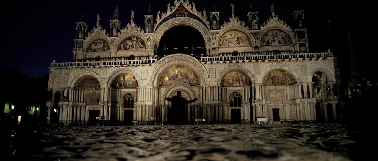 Youth (2015) movie still: wide — A large stone basilica facade with intricate carvings and mosaics glows against the night sky. A person stands in a…