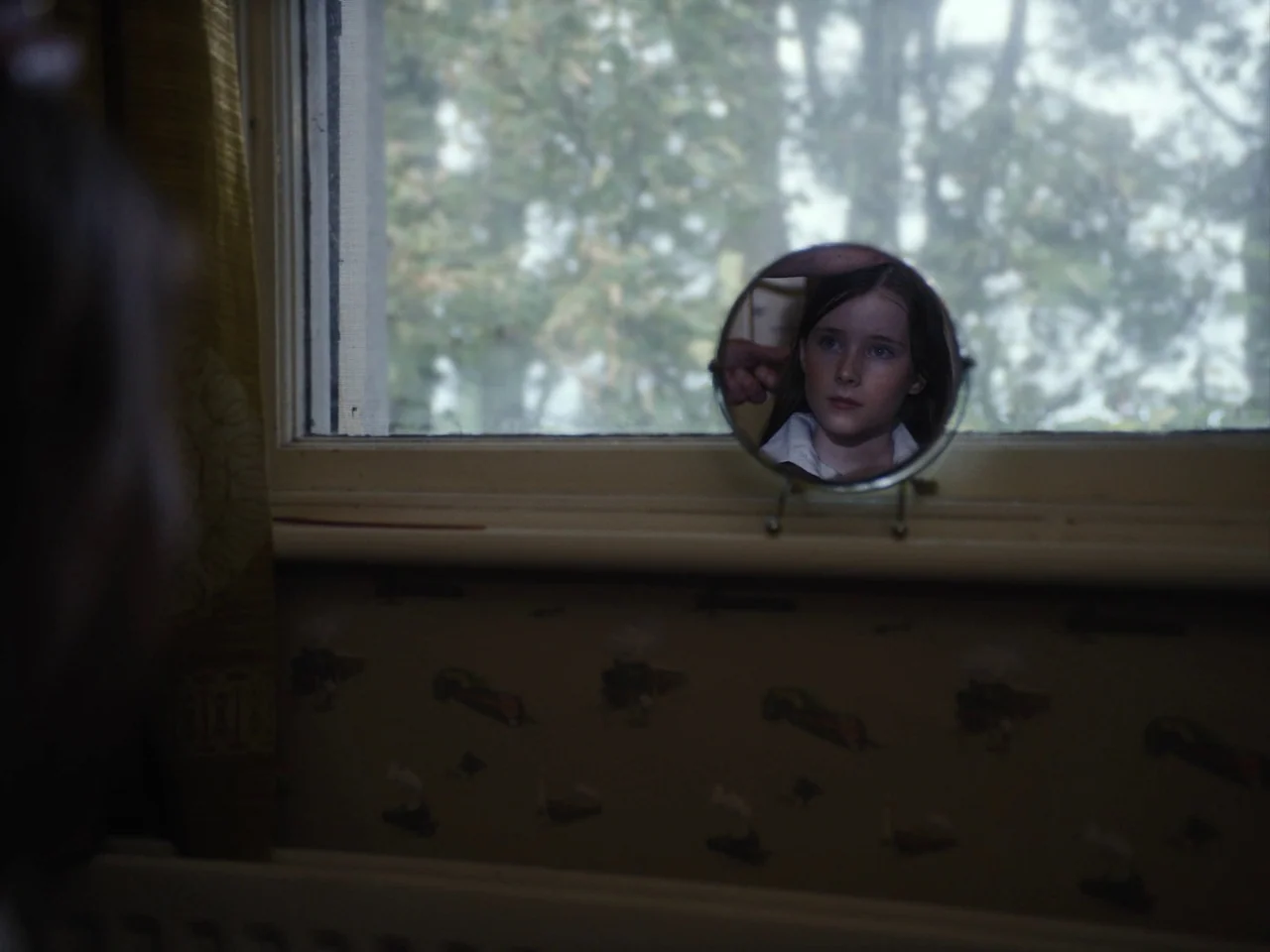 The Quiet Girl (2022) movie still: medium — A child's face is seen reflected in a small circular vanity mirror placed on a windowsill. The…