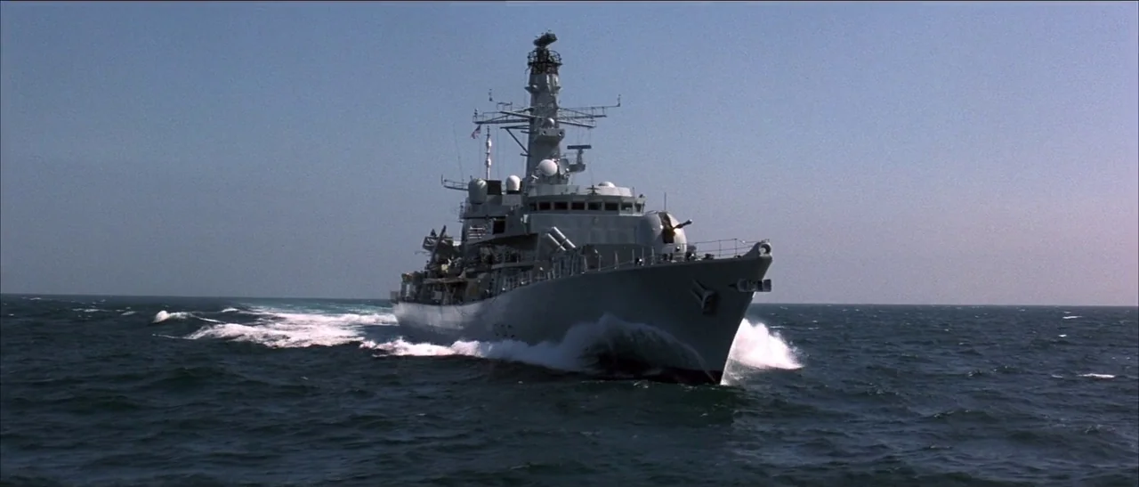 Tomorrow Never Dies (1997) movie still: wide — A grey naval destroyer moves through open dark blue water on a clear day. White spray breaks from the…