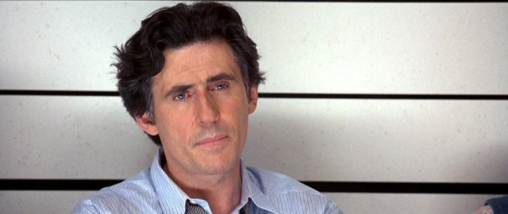 The Usual Suspects movie still: closeup — A man stands before a white wall with horizontal black stripes used for identification. He looks forward…