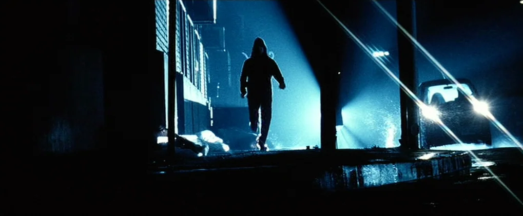 Minority Report movie still: fullbody — A dark urban environment with wet asphalt surfaces and a brick wall. A silhouetted figure in a hoodie walks…