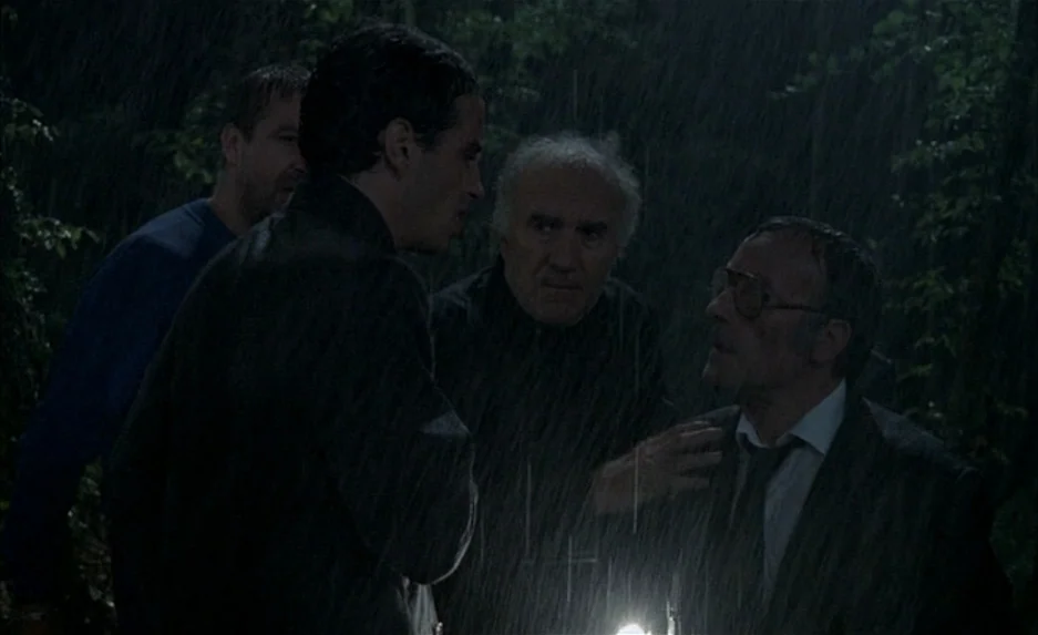 Milou en Mai (1990) movie still: medium — Four men stand close together in a dark, rain-drenched outdoor setting at night. They appear to be in a…