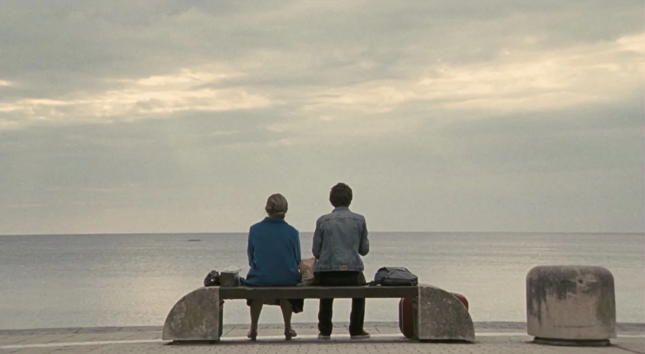 Patagonia (2010) movie still: wide — A stone bench sits on a paved seaside promenade overlooking an expansive, calm gray ocean. A woman and a young…