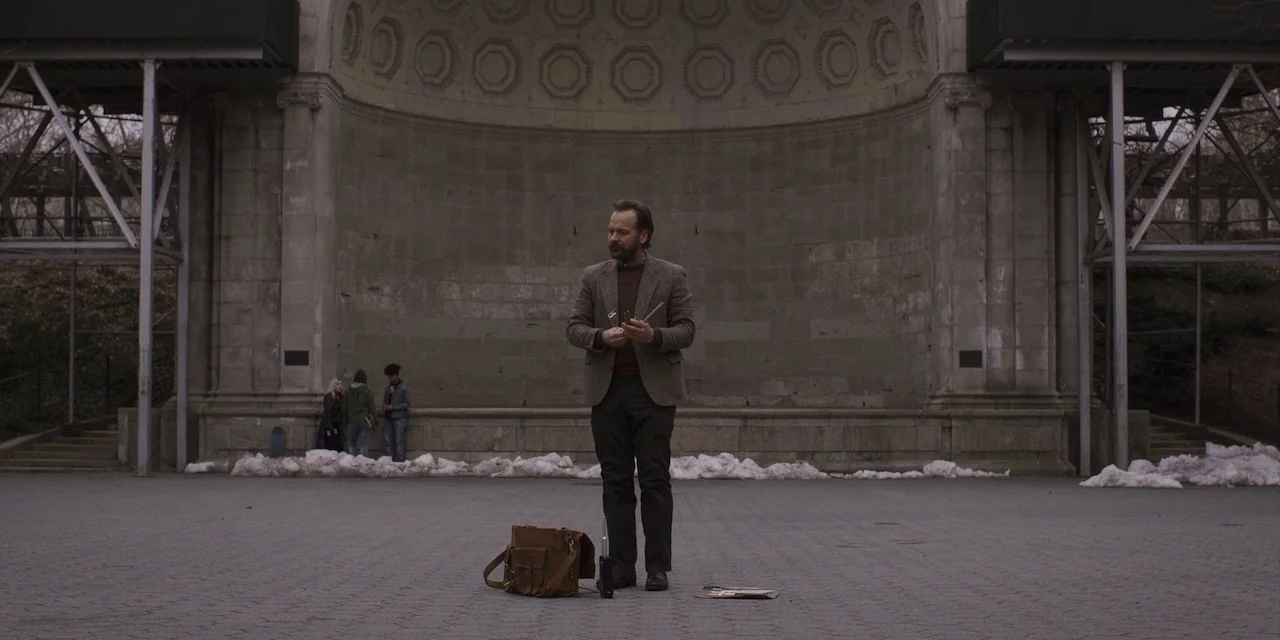 The Sound of Silence movie still: fullbody — A stone bandstand with an arched alcove features a paved plaza in front. A man stands in the center…