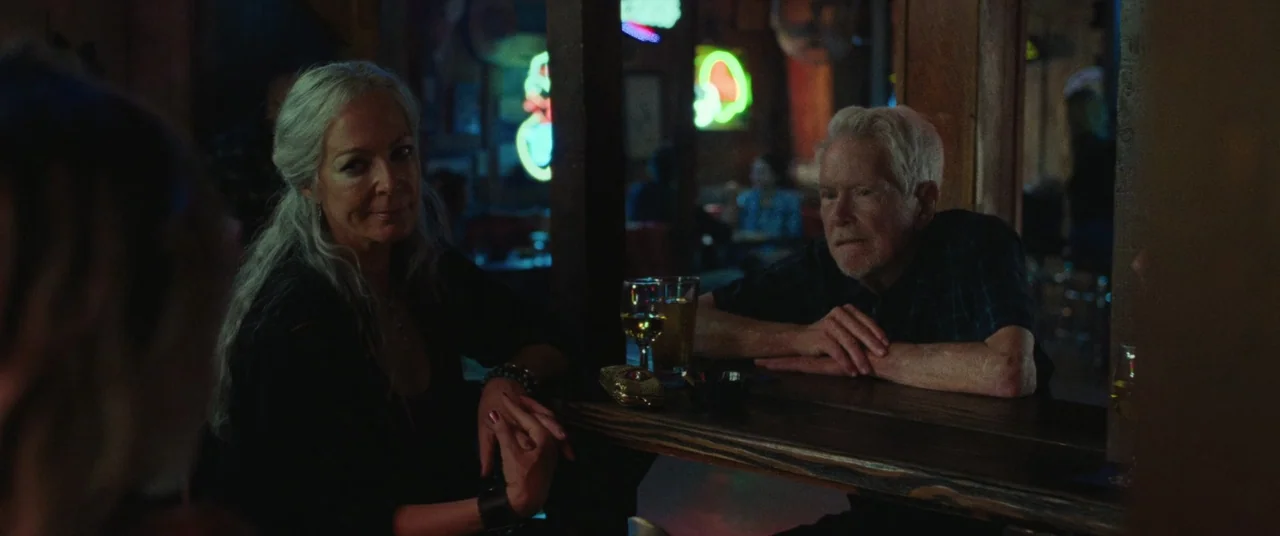 To Leslie (2022) movie still: medium — A dimly lit establishment with dark wood furniture and glowing neon signs in the background. An elderly man…