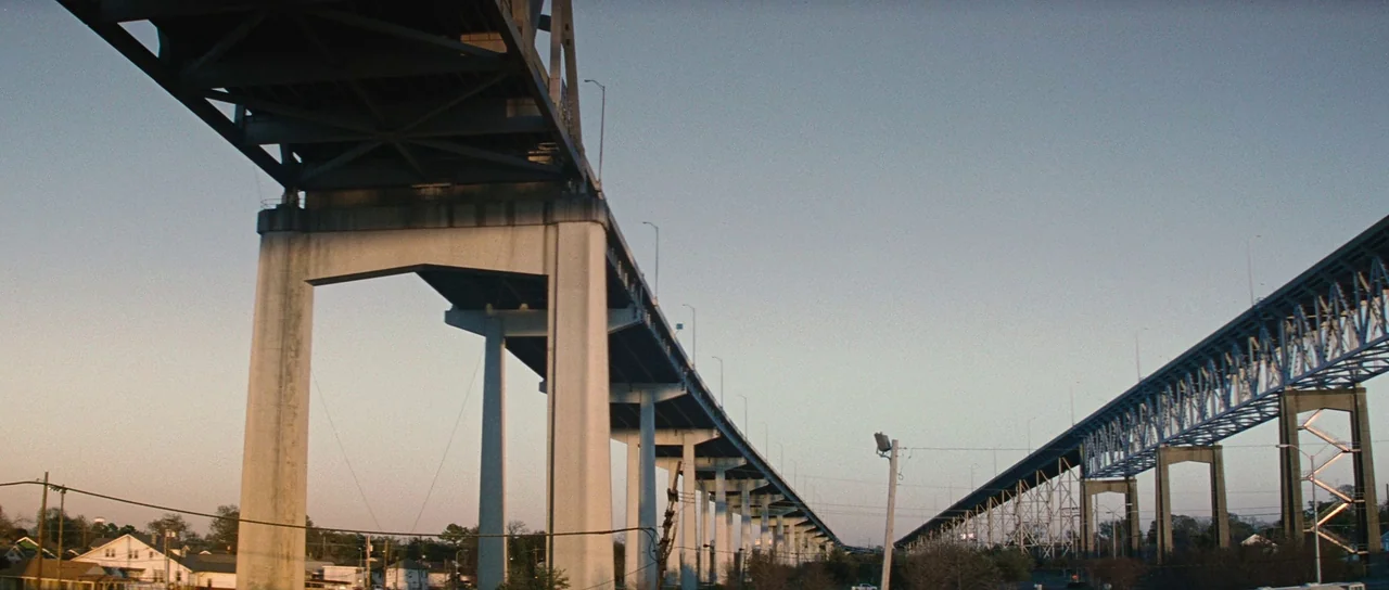 Looper (2012) movie still: establishing — A high-clearance concrete highway bridge spans over a residential area at golden hour. The structures…