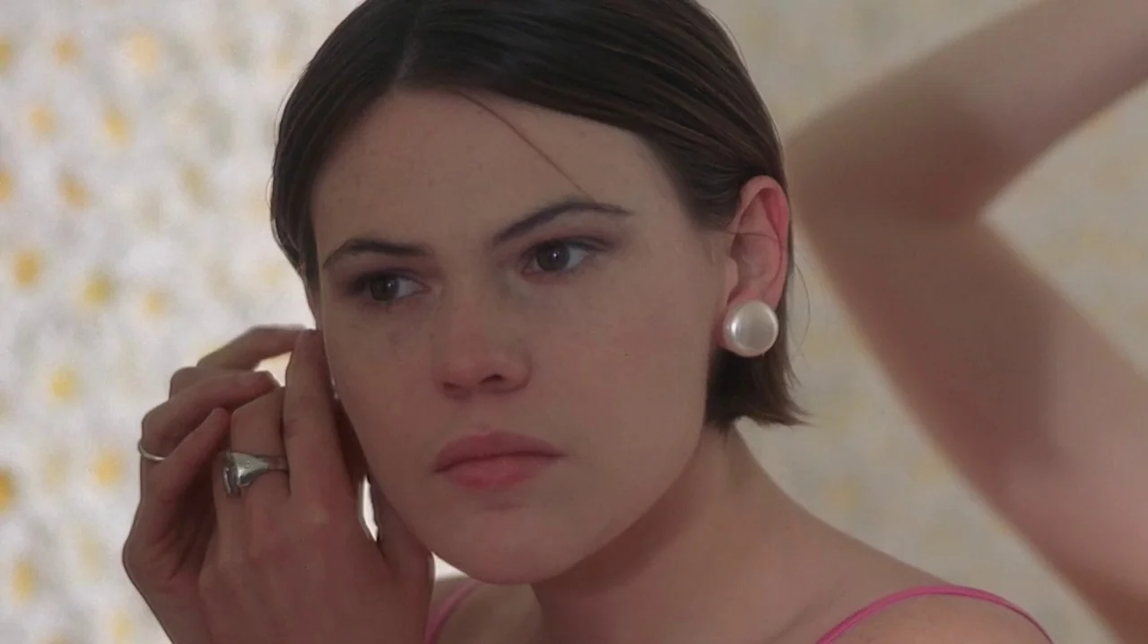 But I’m A Cheerleader (1999) movie still: closeup — A woman stares into a mirror against a background of daisy-patterned wallpaper. She is focused…