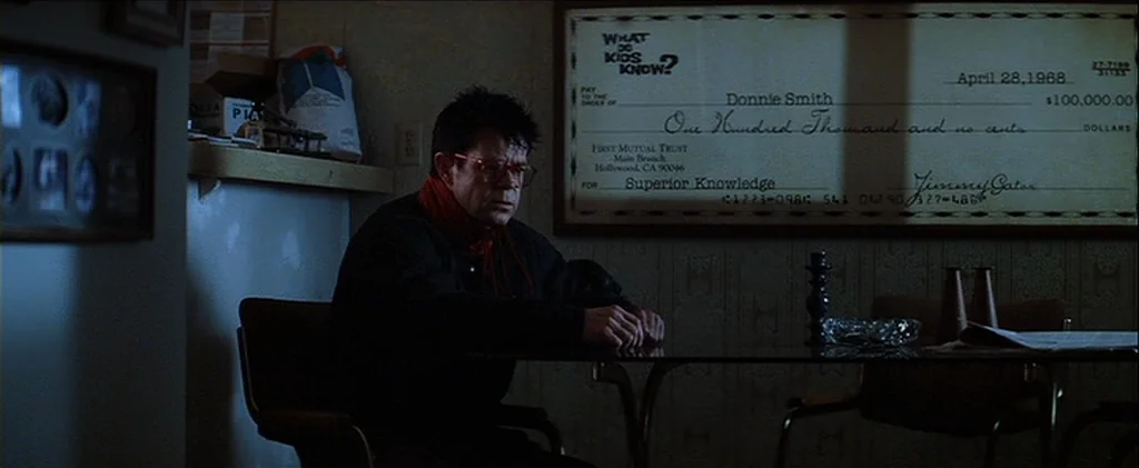 Magnolia (1999) movie still: medium — A dimly lit room features a large framed check hanging prominently on the wall. A man in dark clothing sits at…
