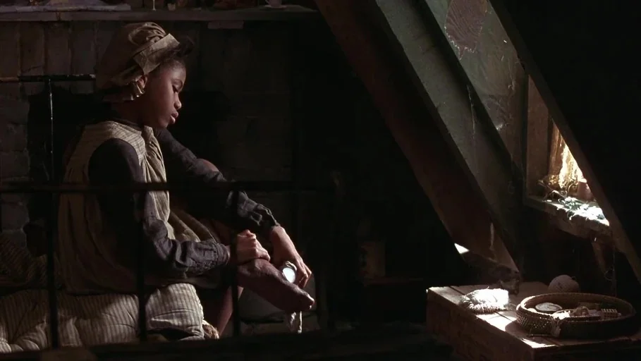 A Little Princess (1995) movie still: medium — A dimly lit attic room with slanted walls and wooden surfaces. A young woman sits on a bed applying a…
