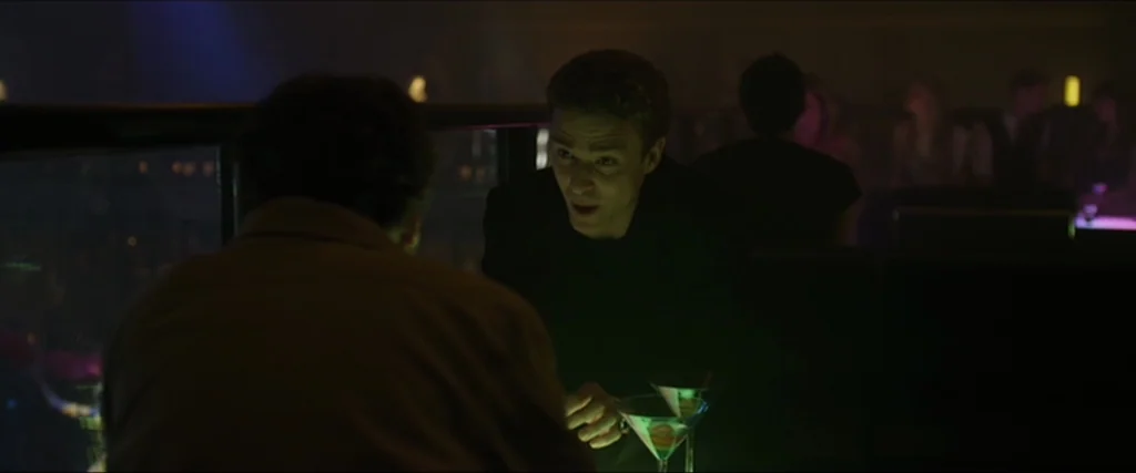 The Social Network movie still: medium — A dimly lit lounge setting features low tables and blurred figures in the background. A man leans forward…