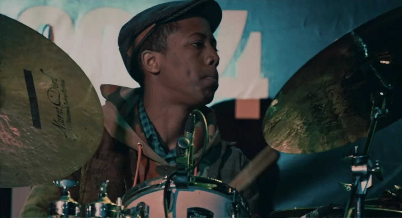 Cairo Jazzman (2017) movie still: medium — A dimly lit indoor space features a drum kit setup against a wall. A young man sits at the drums and…