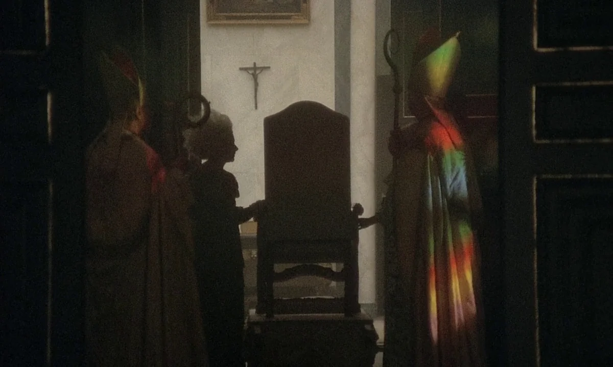 Immoral Tales (1974) movie still: medium — The interior of a church with a crucifix hanging on a white wall. Two bishops stand by a wooden chair as…
