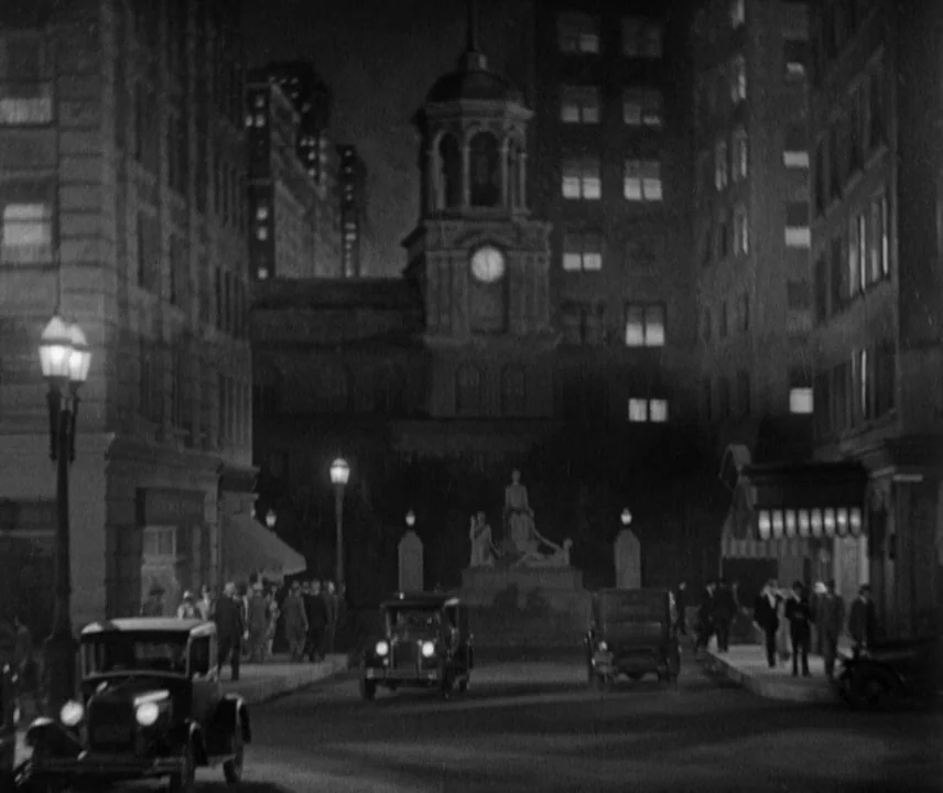 City Lights (1931) movie still: establishing — A city street at night featuring a clock tower and a central statue. Vintage automobiles drive along…