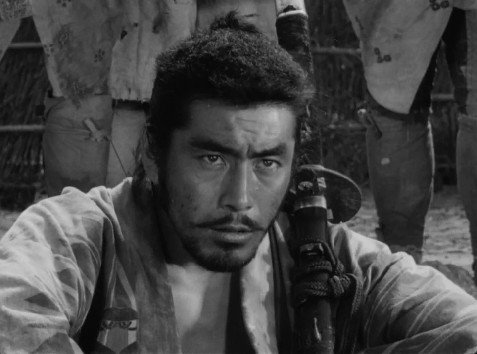Seven Samurai (1954) movie still: closeup — A man in traditional attire crouches in an outdoor environment with legs visible behind. He stares…