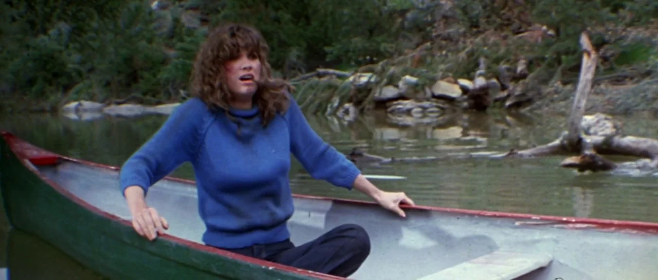Friday the 13th Part III (1982) movie still: medium — A woman sits in a green and red canoe on calm water near a shoreline. Behind the canoe,…