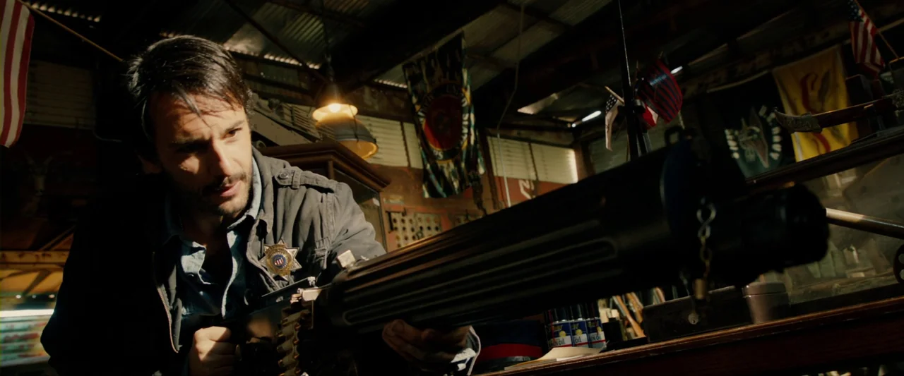 The Last Stand (2013) movie still: medium — A cluttered shop interior features military flags hanging from the corrugated metal ceiling rafters. A…