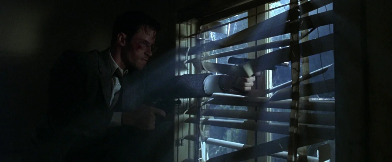 L.A. Confidential (1997) movie still: medium — A dark, spartan room features a window covered by horizontal blinds at night. A man in a suit…