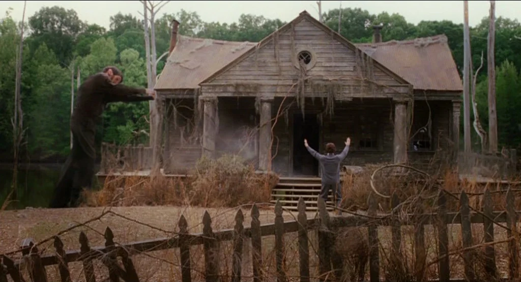 Big Fish (2003) movie still: wide — A weathered wooden house sits on the edge of a still body of water. A man stands in the yard with his arms…