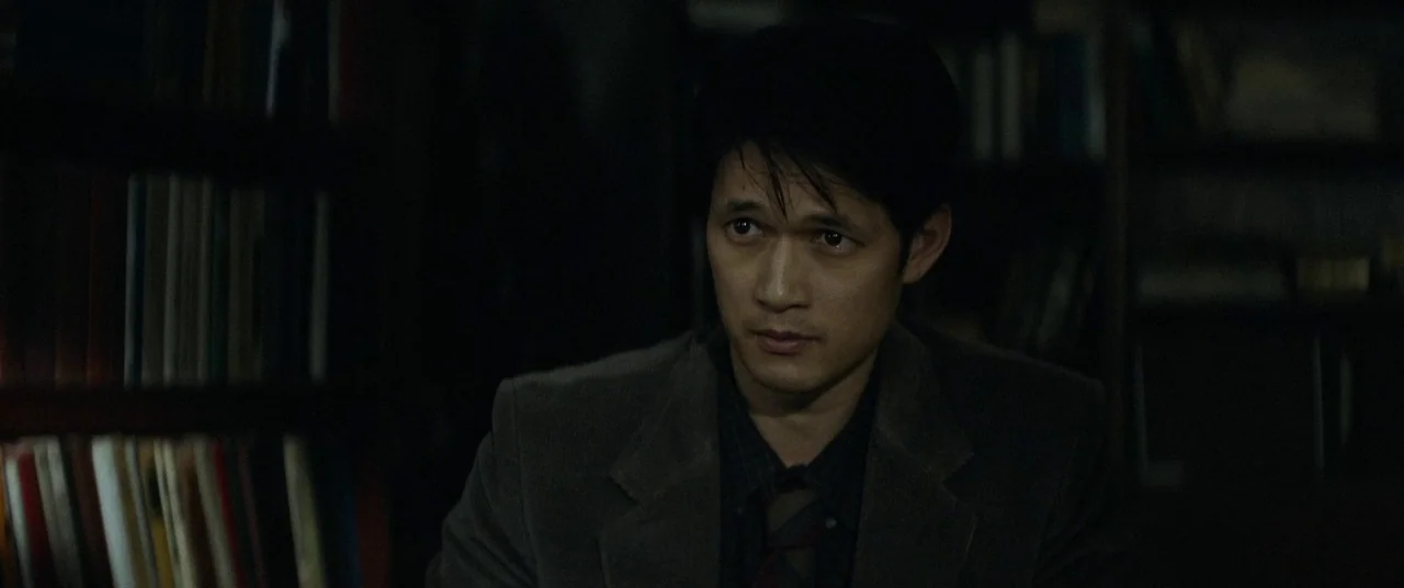 Broadcast Signal Intrusion (2021) movie still: medium — A dark, dimly lit library with shelves filled with books in the background. A man sits in…