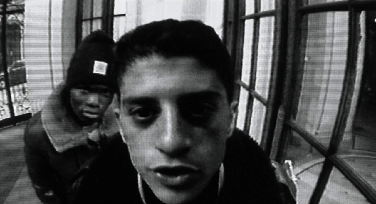 La Haine (1995) movie still: closeup — Two men stand directly in front of a fisheye security camera at a building entrance. The footage appears to…