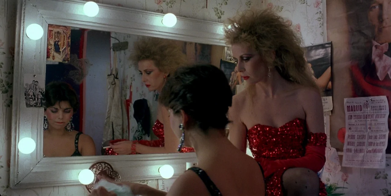 Dark Habits (1983) movie still: medium — A cluttered backstage dressing room with floral wallpaper and posters of theatrical events. Two women…